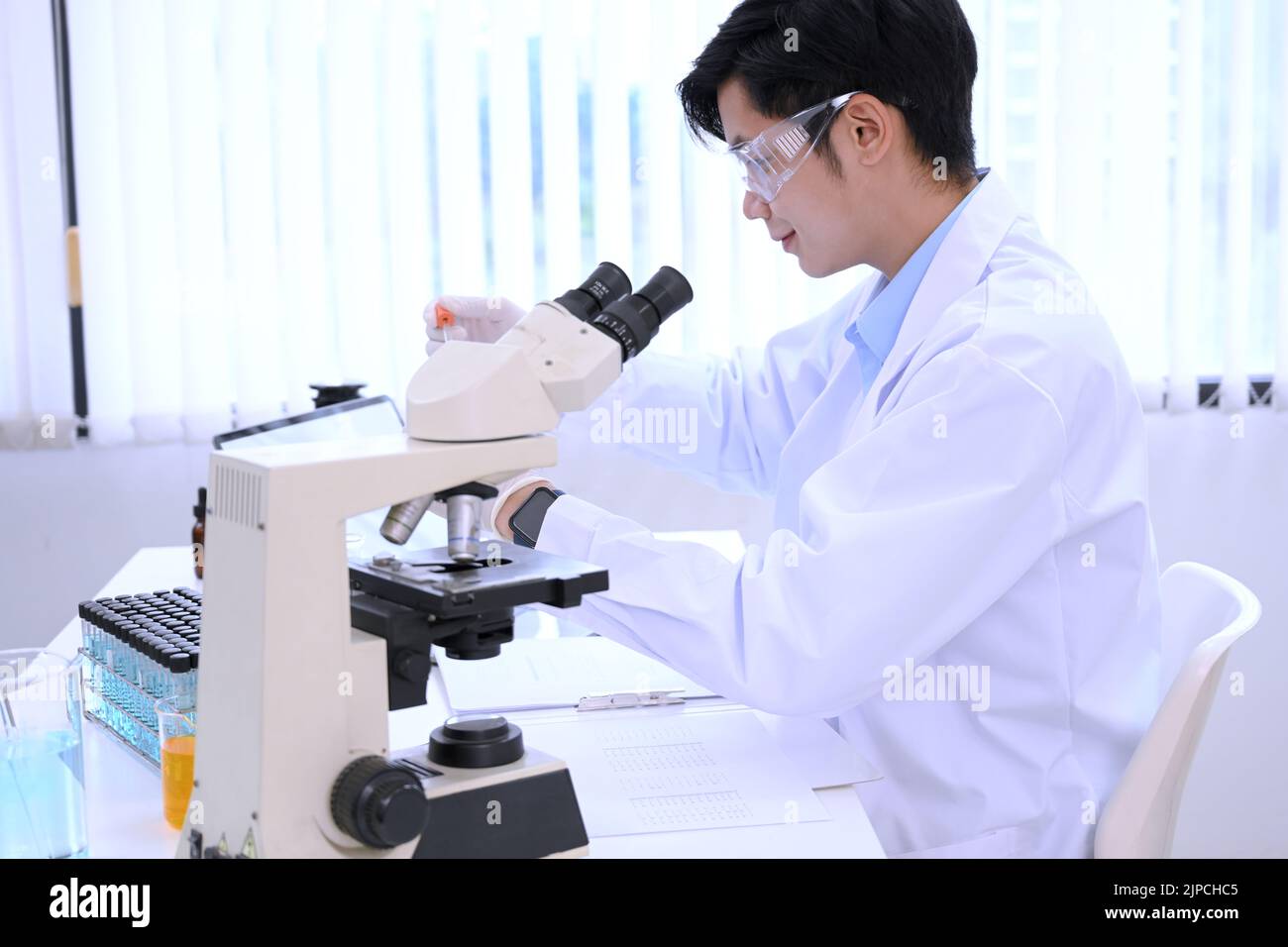 Giovane scienziato maschile asiatico concentrato o specialista che lavora nel suo laboratorio d'ufficio, lavorando sul microscopio e sulla carta di ricerca. Foto Stock