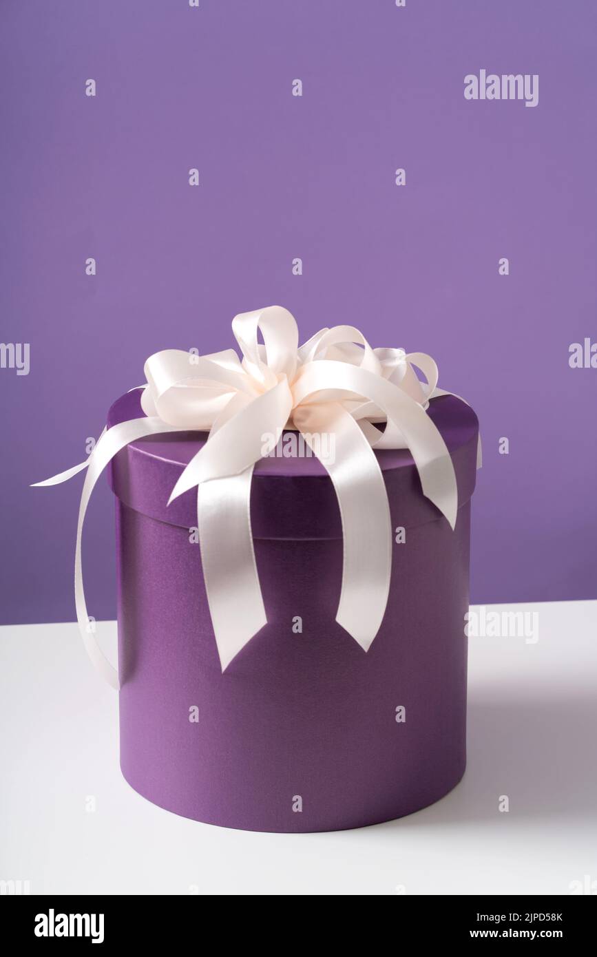 Una scatola rotonda viola con un arco bianco soffuso su un tavolo bianco. Concetto di vacanza. Foto Stock