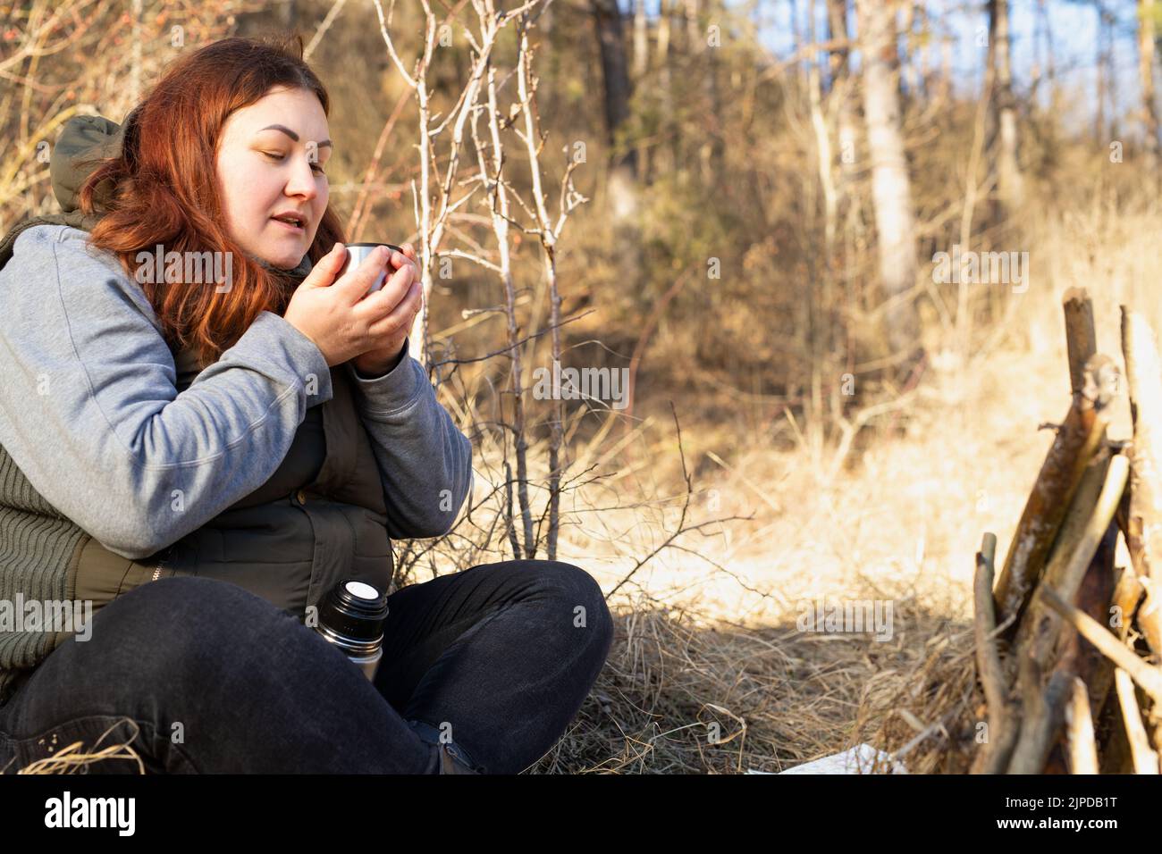 Viaggiatore donna grassa che indossa abiti casual seduto con gambe incrociate nella foresta vicino al falò tenendo in mano una tazza di tè caldo. Foto Stock