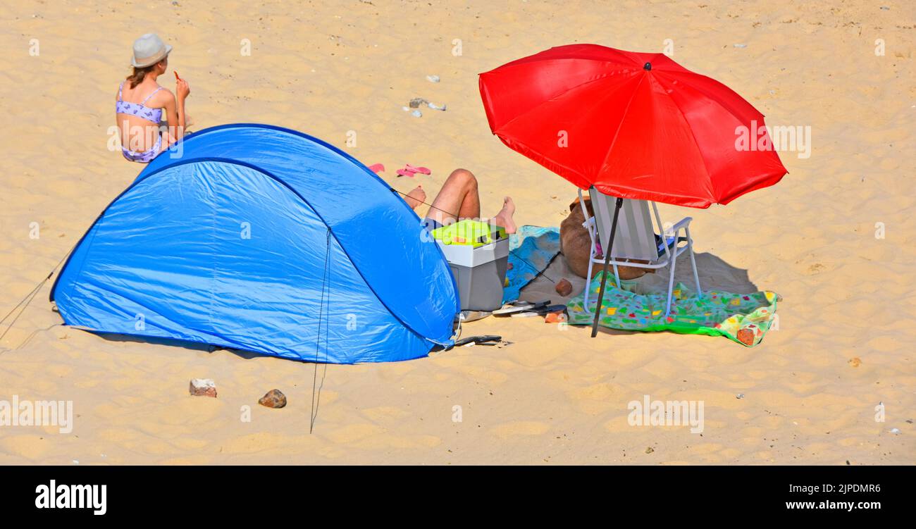 Coppia con il cane dell'animale domestico all'ombra sulla spiaggia sabbiosa di mare che ripartisce il parasole rosso colorful ombrellone & tenda polivalente blu in vacanza soleggiata calda del Regno Unito di luglio Foto Stock