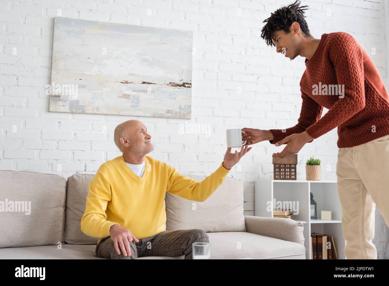 Vista laterale del nipote afro-americano che dà la tazza al nonno sorridente a casa Foto Stock