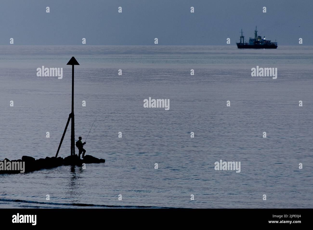 Una silhouette non identificabile di un uomo che pesca alla fine di un molo con la silhouette non identificabile di una nave all'orizzonte. Foto Stock