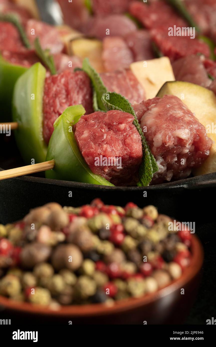 Macro primo piano di spiedini di carne cruda prima della cottura su fondo scuro con foglie di salvia e pepe misto. Foto Stock