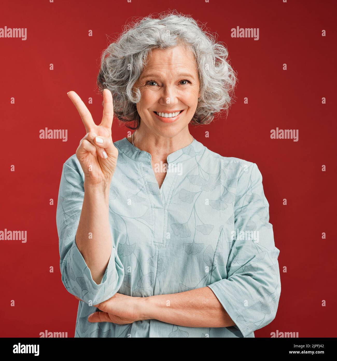 .. Donna anziana esprime segno di pace, v gesto ed emoji con le dita su sfondo rosso studio. Ritratto di donna spensierata, fresca e sorridente in positivo Foto Stock