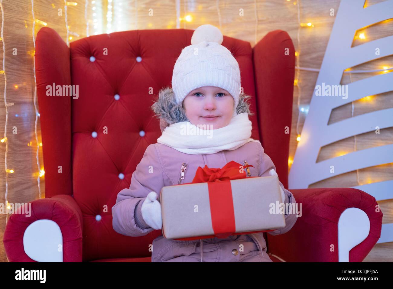 Ritratto piccola ragazza che si siede in una sedia rossa di natale e tiene il regalo di Natale all'esterno. Natale presenta il concetto di infanzia di Vacanze Foto Stock