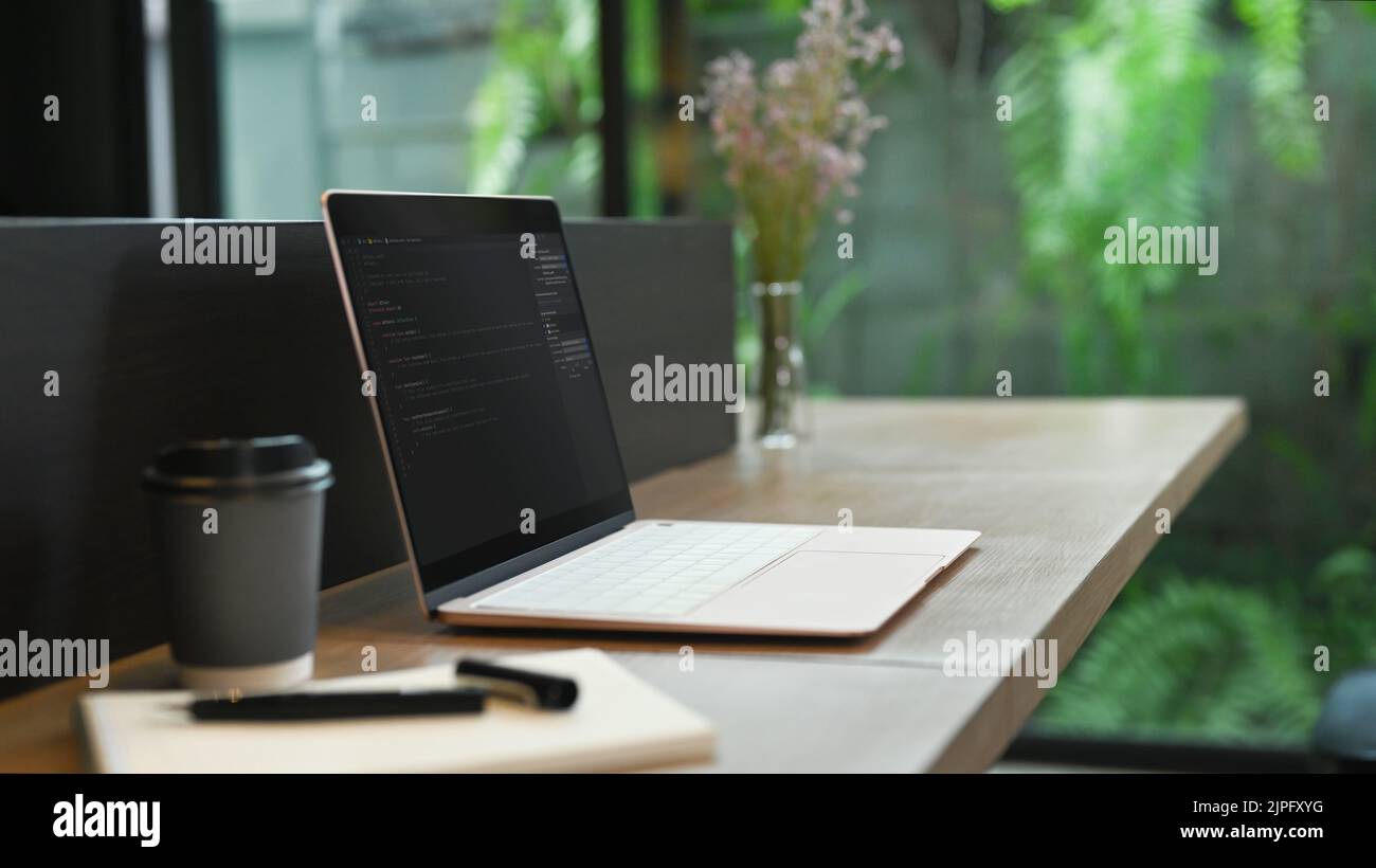 Computer portatile con codice di programmazione sullo schermo, tazza da caffè e notebook su scrivania in legno Foto Stock