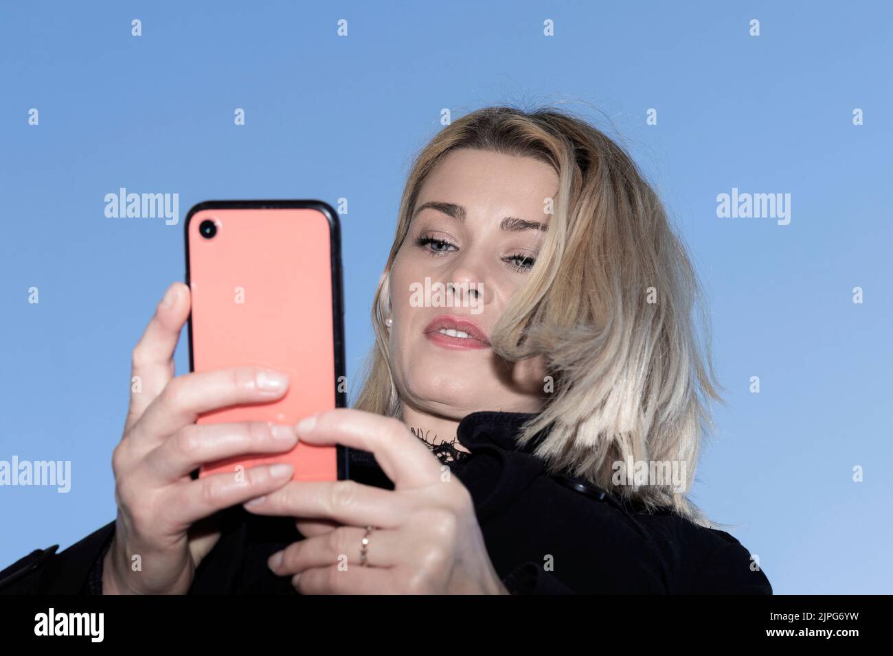bella donna bionda guardando un telefono cellulare rosso con il cielo dietro Foto Stock