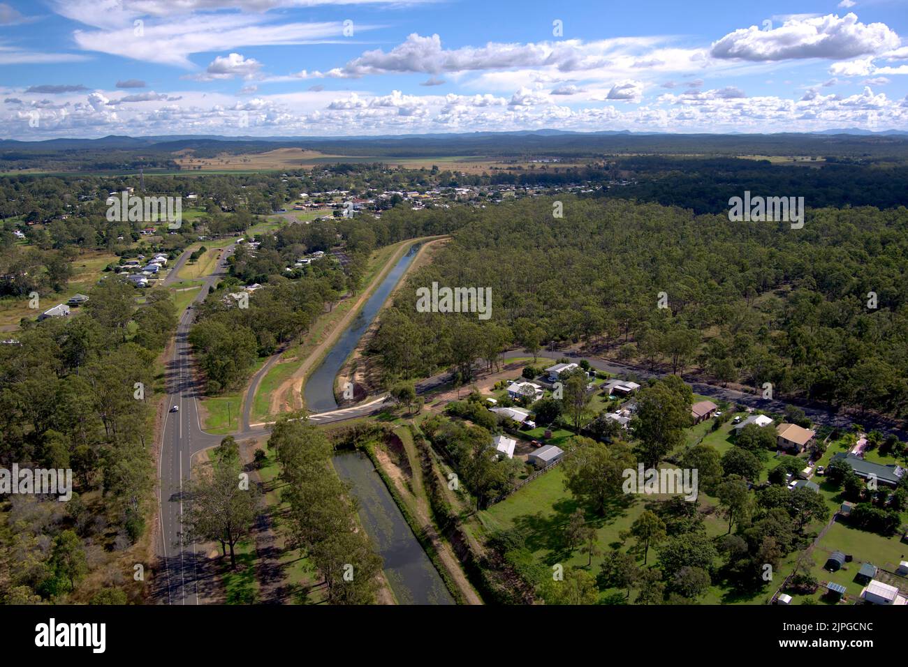 Vista aerea di una strada curva e di un canale di irrigazione attraverso un paesaggio suburbano con case e vegetazione sotto un cielo blu con nuvole Foto Stock