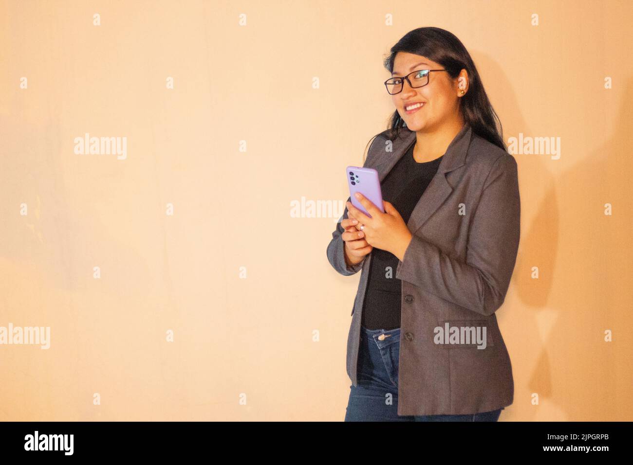 Una donna caucasica che indossa una giacca grigia che tiene il telefono e guarda la fotocamera con un sorriso Foto Stock