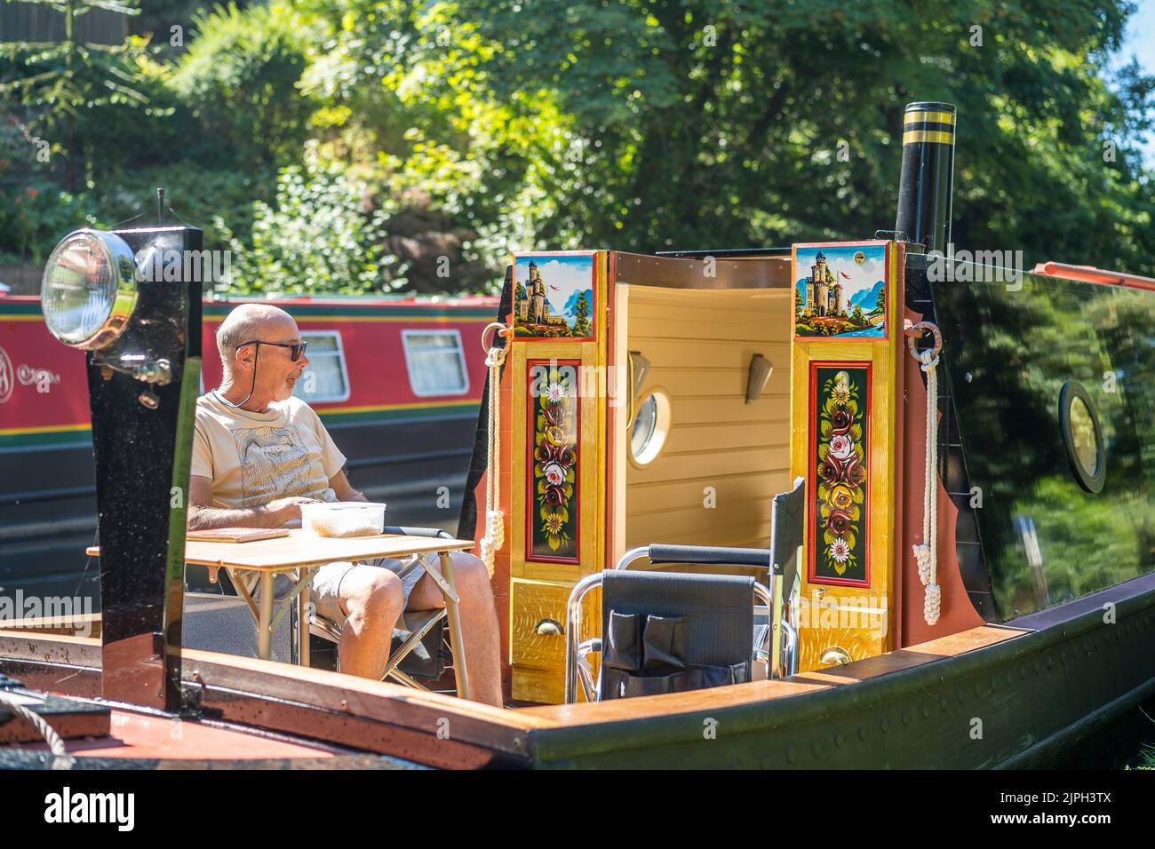 UK narrowboat ormeggiato a lato del canale Worcestershire (le porte si aprono mostrando rose e castelli tradizionali disegno di vernice) mentre l'uomo si siede godendo l'onda di calore. Foto Stock