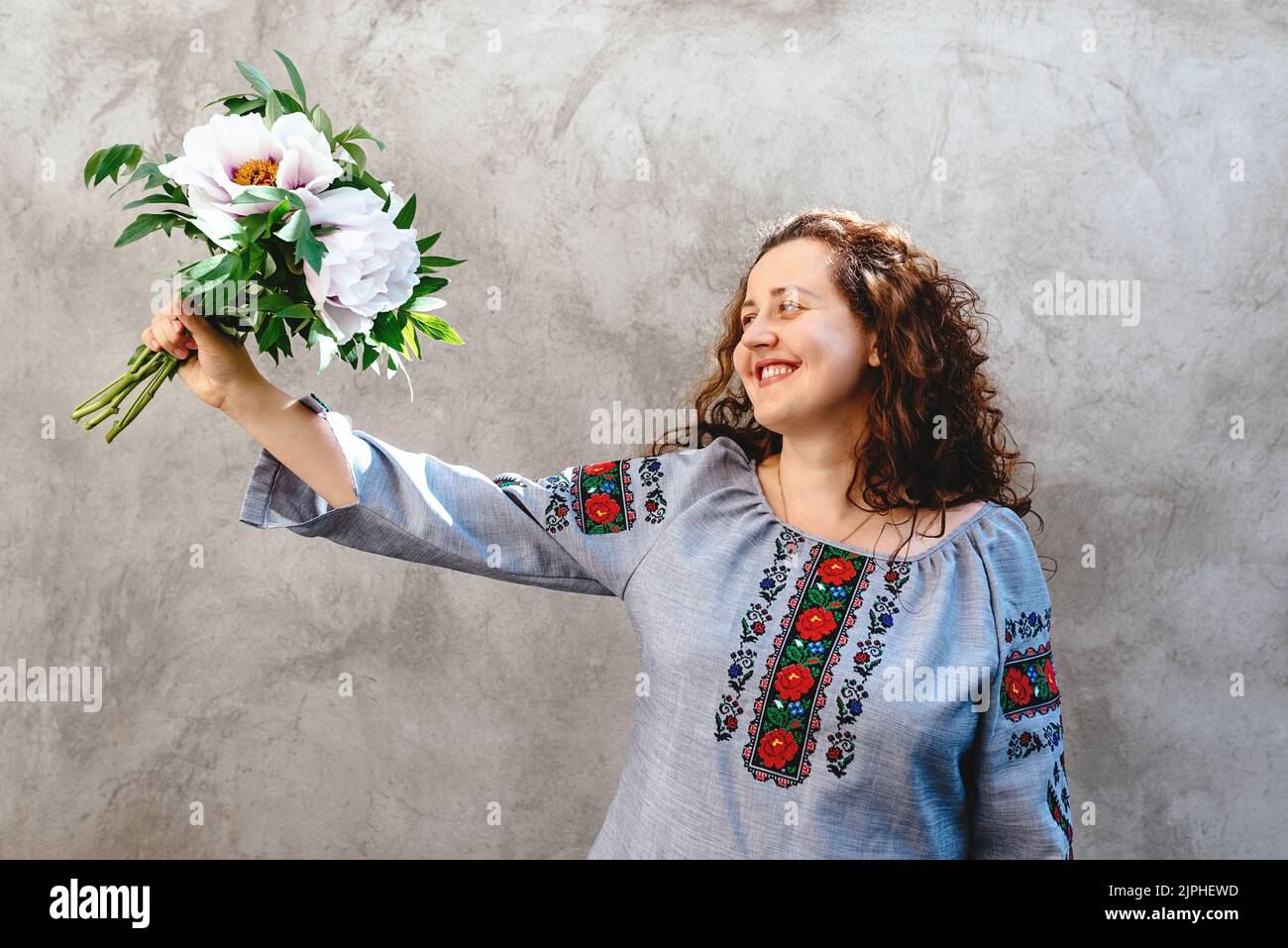 La ragazza sorridente sta tenendo il bouquet delle peonies rosa. La donna indossa una tradizionale camicia Ucraina ricamata sullo sfondo di una parete di cemento. Giornata dell'indipendenza dell'Ucraina. Spazio di copia. Messa a fuoco morbida Foto Stock