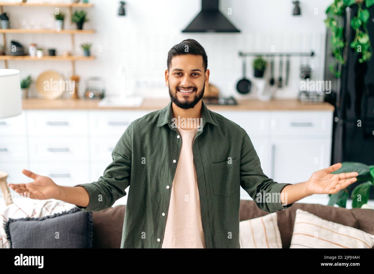 Felice positivo moderno indiano o arabo ragazzo, in abiti casual elegante, in piedi a casa nel soggiorno sullo sfondo della cucina, si distende le braccia a lato, guarda la macchina fotografica, sorriso Foto Stock