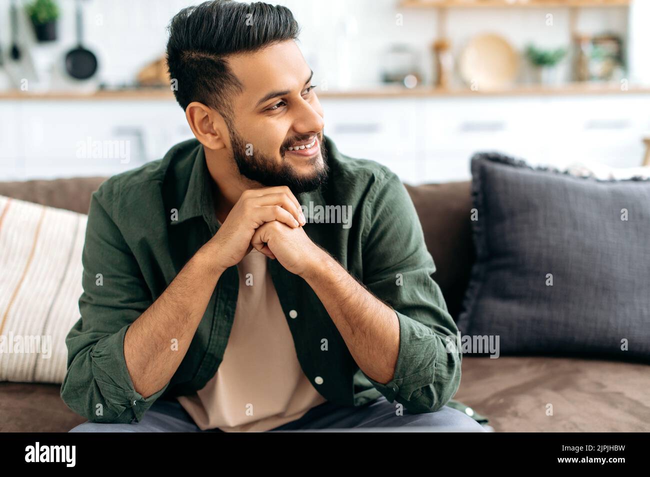 Bel positivo sicuro attraente indiano o arabo uomo, in abiti casual elegante, seduto a casa in accogliente soggiorno sullo sfondo della cucina, guarda a lato, sorridendo Foto Stock