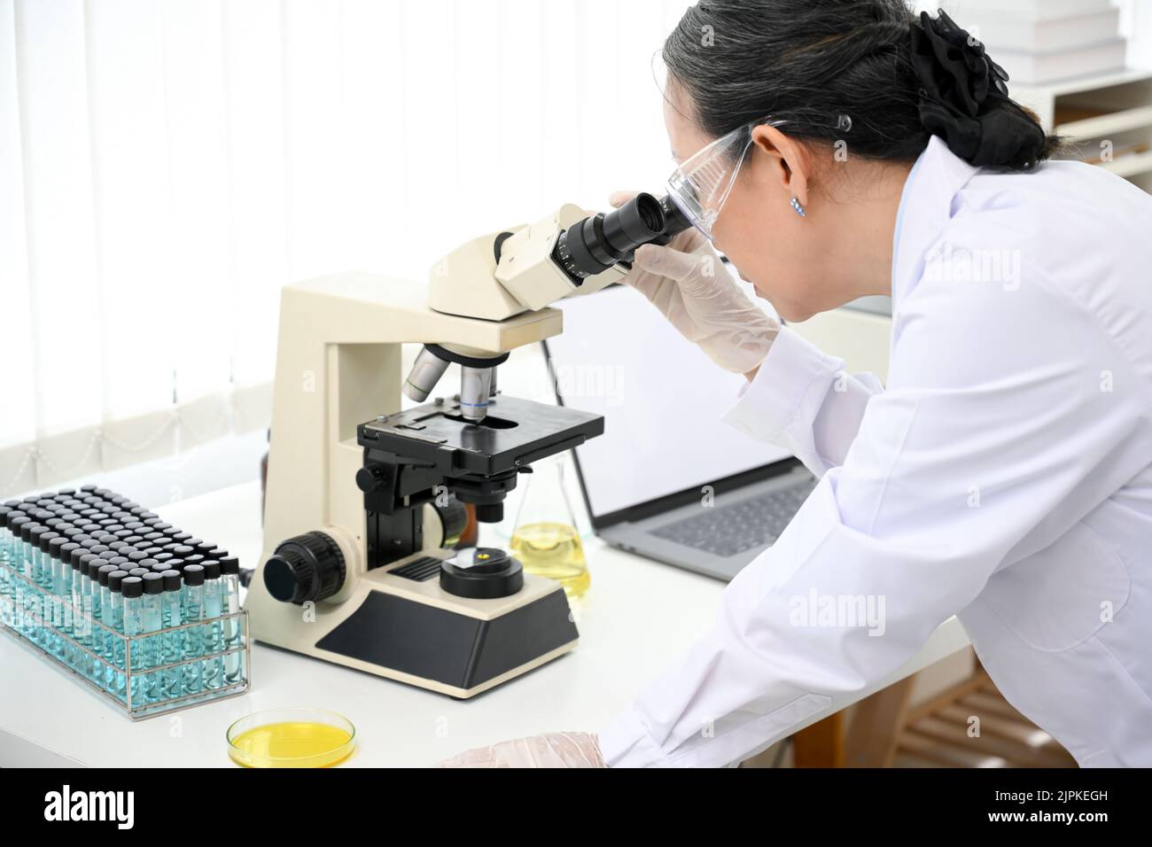 Un esperto scienziato asiatico di età femminile o un supervisore chimico medico che esamina un campione di virus sotto un microscopio mentre lavora in laboratorio. Primo piano i Foto Stock