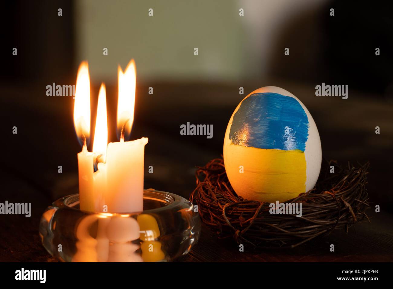 Un uovo di pollo bianco con una bandiera dipinta dell'Ucraina in un cesto di paglia su un tavolo a casa con una fiamma di candela, cultura e vacanze 2022 Foto Stock