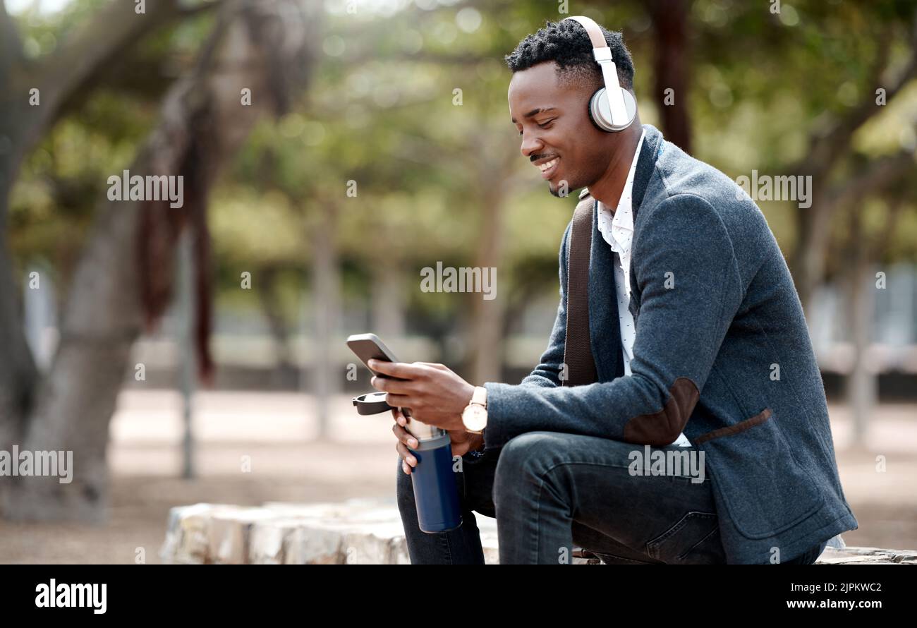 Un uomo d'affari che naviga sui social media sul telefono e che si diverti con la sua app per playlist musicali in cuffie all'aperto. Giovane, felice e casual africano maschio Foto Stock