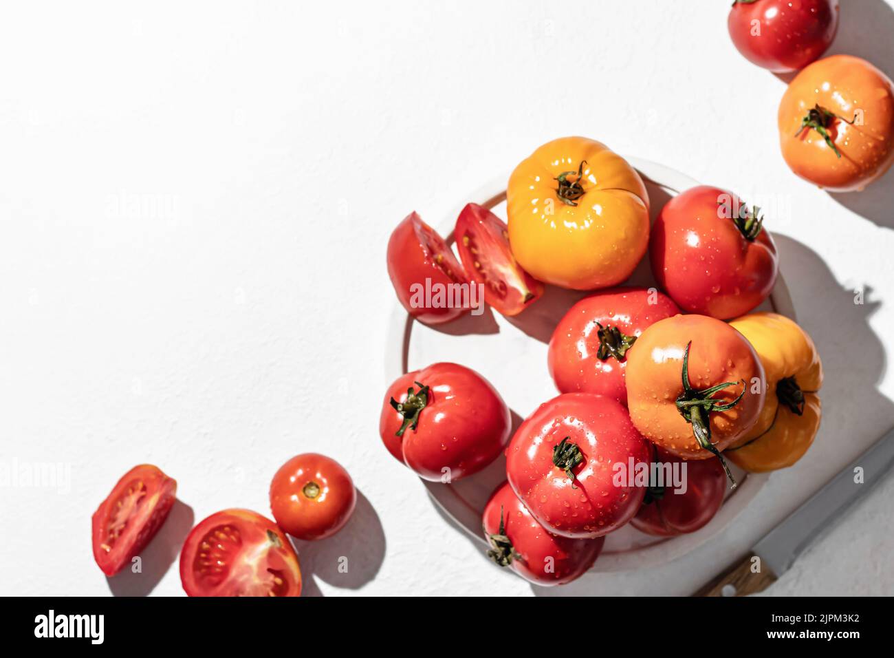 Pomodori freschi e colorati e coltello sul piatto con ombre dure su sfondo bianco con spazio copia, vista dall'alto Foto Stock