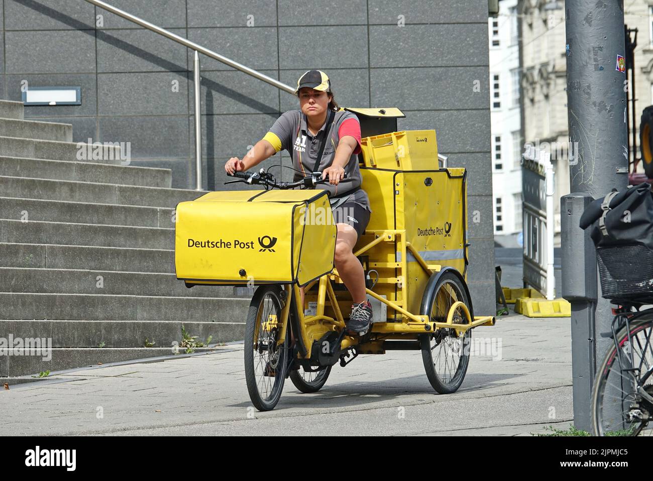 Postmistress a bordo della sua bici da carico che trasporta la posta. Posta consegna pacchi in Germania. Monaco di Baviera, GERMANIA - Agosto 2022 Foto Stock