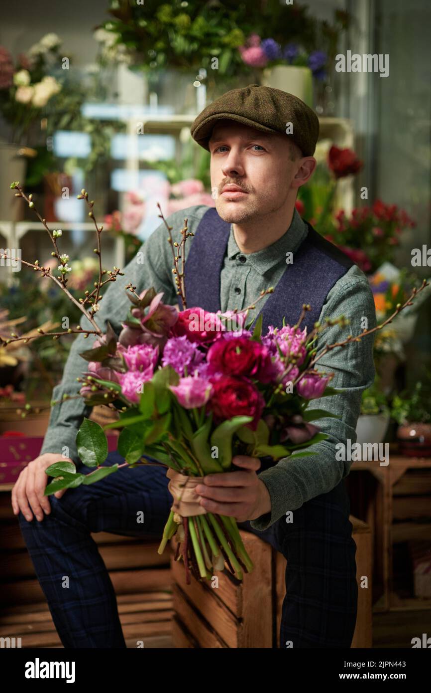 Maschio caucasico in berretto e vintage vestiti dal 20s tenendo bel bouquet di fiori rossi e violetti in negozio di fiori. Congratulazioni con i fiori. Il giorno della mamma o il giorno di San Valentino Foto Stock