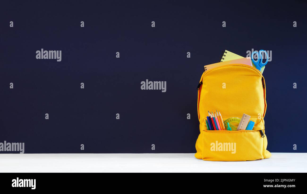 Zaino con diversi coloratissimi articoli da cancelleria sul tavolo e sfondo blu. Banner design di ritorno a scuola concetto sfondo. Lavagna blu Foto Stock