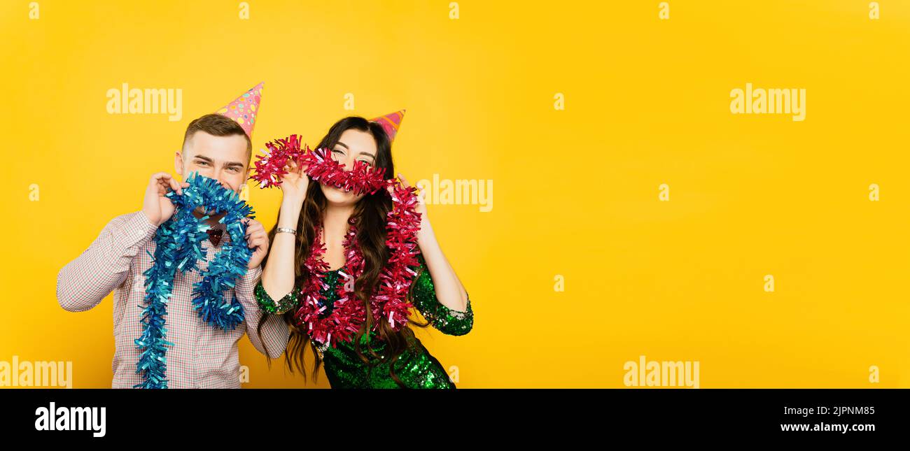 Giovane adulto ragazzo e ragazza 20-25s in cappellino di festa posa con il tinsel di Capodanno, fare baffi e guardare la macchina fotografica su uno sfondo giallo isolato. Fest Foto Stock