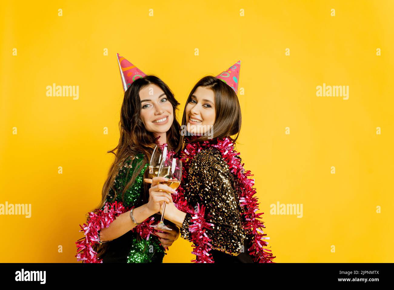 Giovane ragazza allegra 20-25s in cappelli da festa e tinsel di Capodanno, abbracciando bicchieri di vino o champagne e guardando la macchina fotografica su un yel Foto Stock