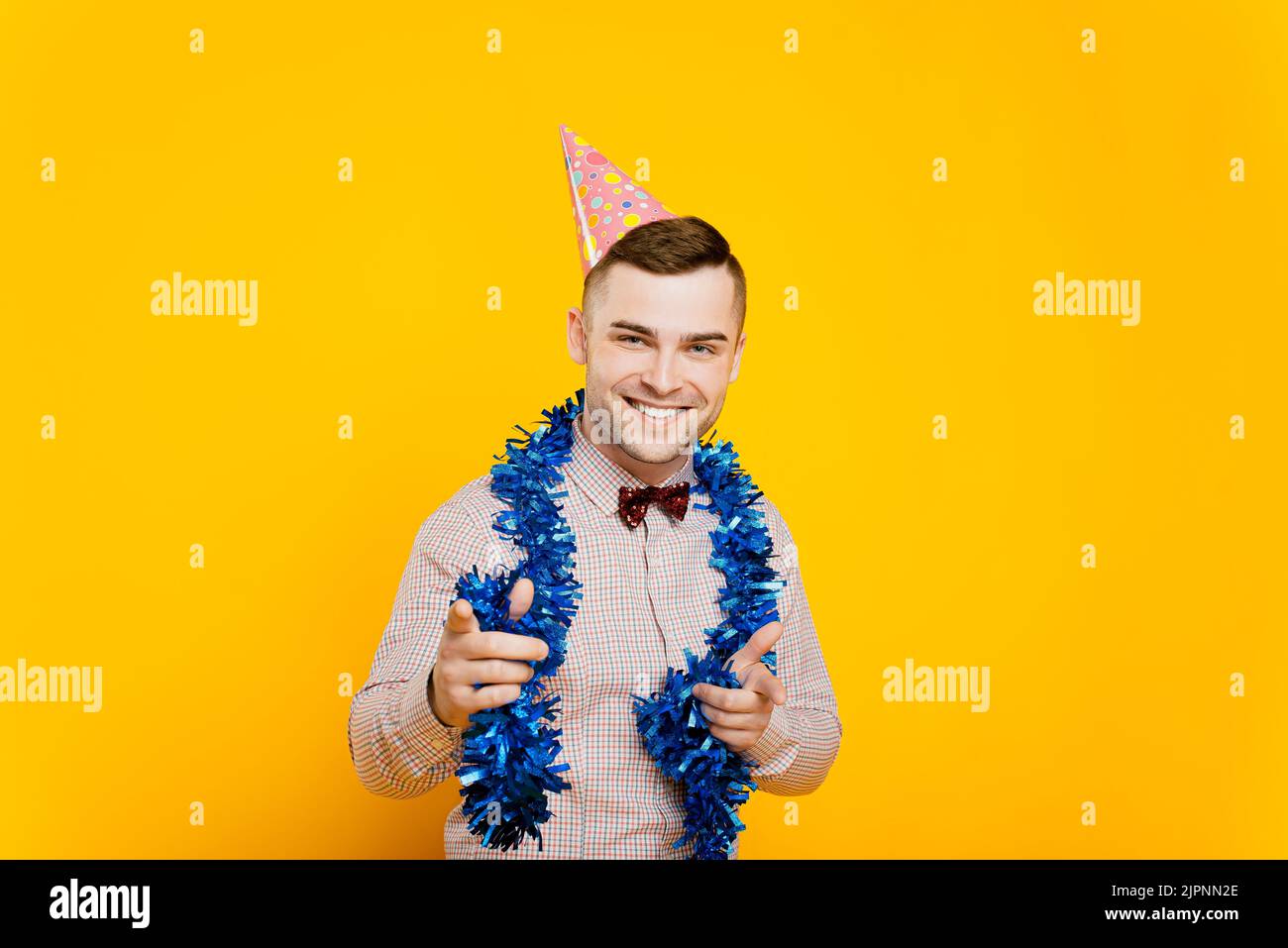 Ragazzo giovane adulto 20-25s con un cappello da festa e tinsel di nuovo anno facendo un gesto che punta con le dita indice in avanti e sorridendo guardando la macchina fotografica Foto Stock