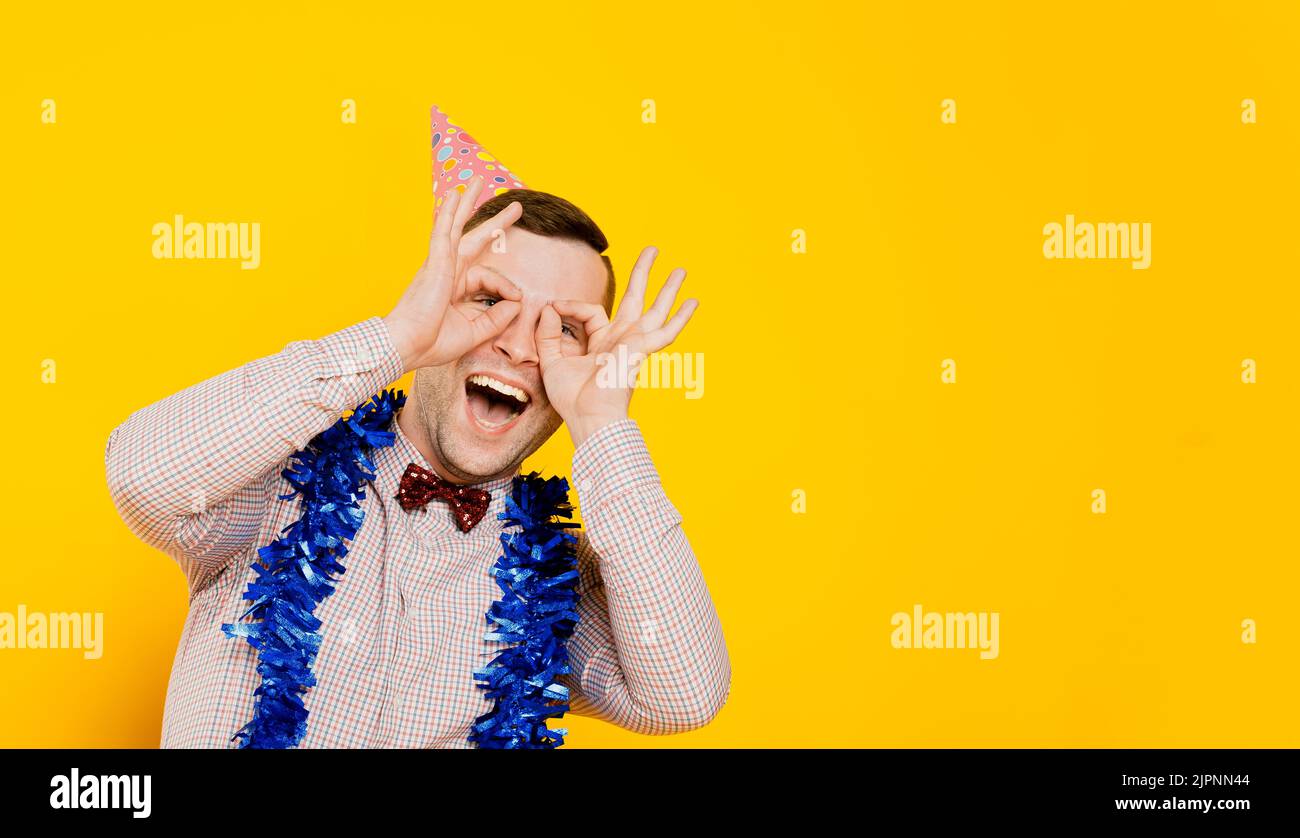 Divertente ragazzo 20-25s in un cappello da festa e il tinsel di Capodanno ride e fa un gesto con le mani sotto forma di occhiali o binocolo, sotto forma di un Foto Stock