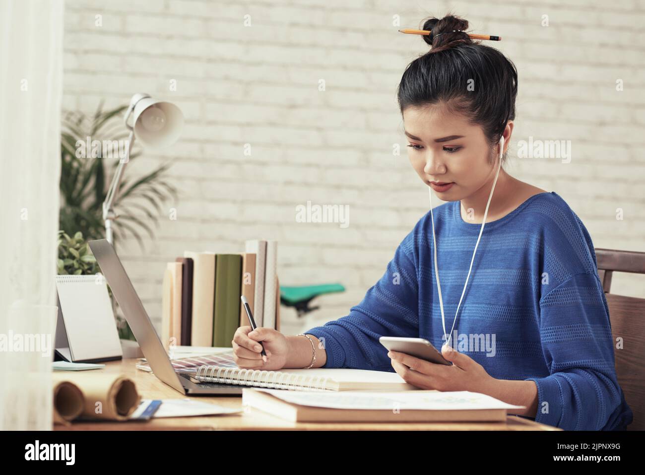 Grazioso studente vietnamita con smartphone che scrive nel suo libro di testo Foto Stock