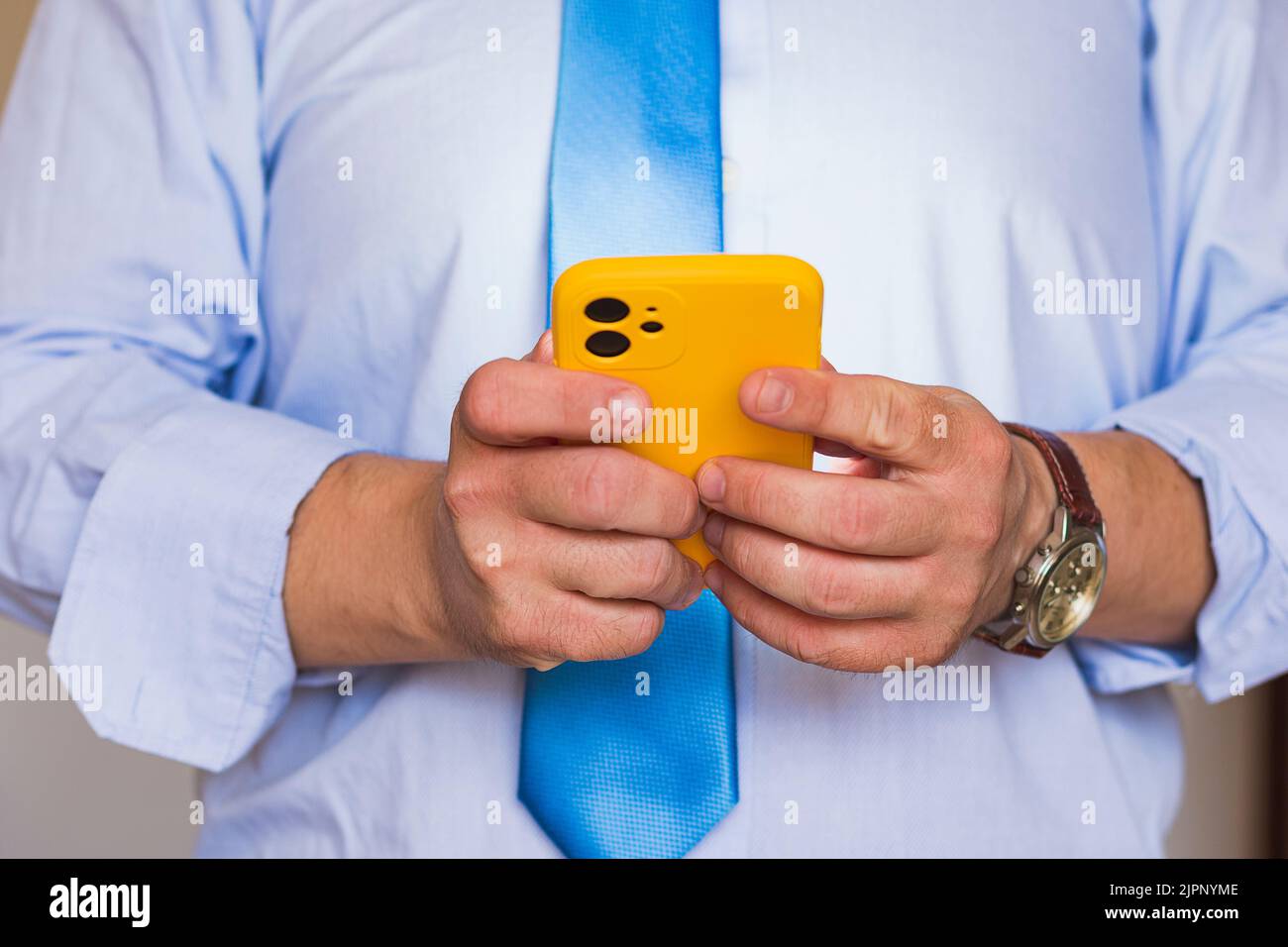 Primo piano delle mani di un uomo caucasico irriconoscibile che tiene in mano un telefono cellulare con una custodia gialla. La persona è vestita con una camicia blu e una cravatta blu, Foto Stock