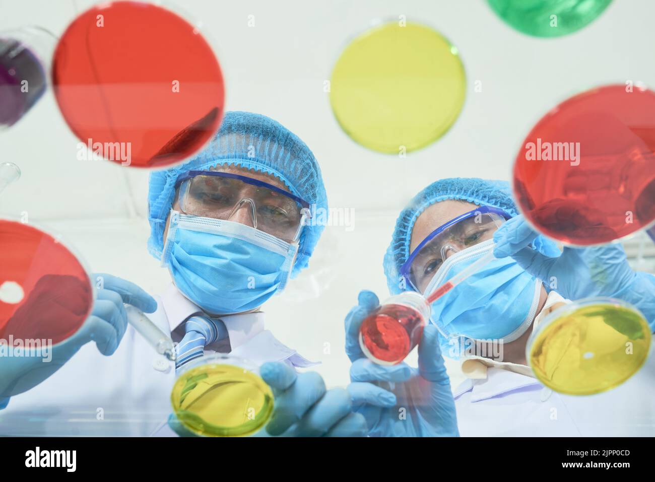 Vista a basso angolo di scienziati asiatici concentrati che indossano camici bianchi e guanti di gomma utilizzando pipette e vetreria da laboratorio durante l'esecuzione di esperimenti, vista attraverso la superficie del tavolo di vetro Foto Stock