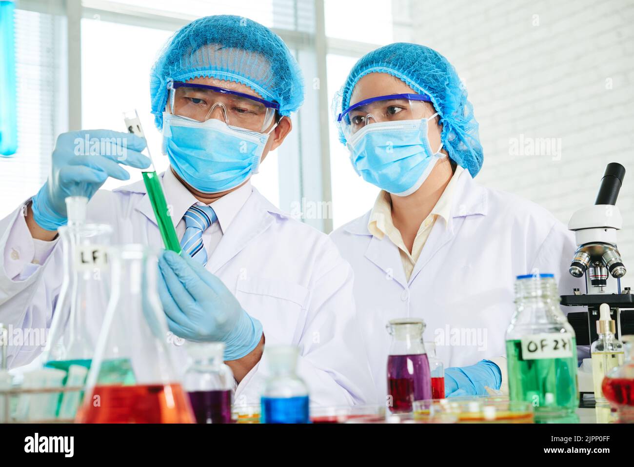 Scienziati asiatici concentrati che indossano camici bianchi e maschere mediche che esaminano il liquido in una provetta mentre sono riuniti in un laboratorio moderno Foto Stock