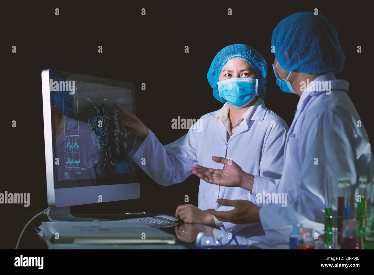 Ricercatori di talento che indossano maschere mediche e camici bianchi che studiano dati statistici mentre si siedono di fronte al computer moderno al laboratorio dim Foto Stock