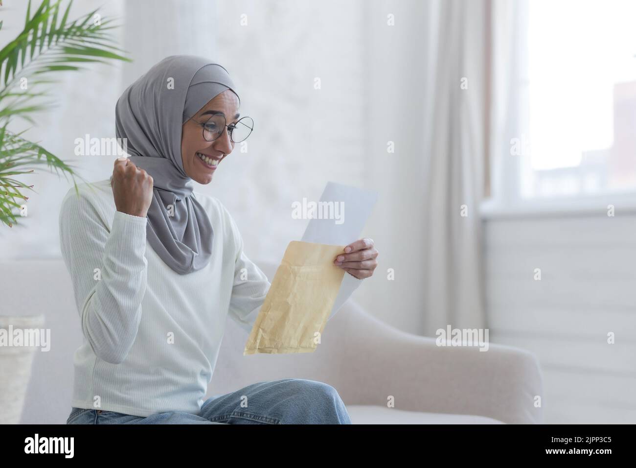 Una giovane bella donna musulmana legge una felice lettera casa studente arabo ha ricevuto conferma dall'università superato con successo l'esame una donna in un hijab è seduta su un divano Foto Stock