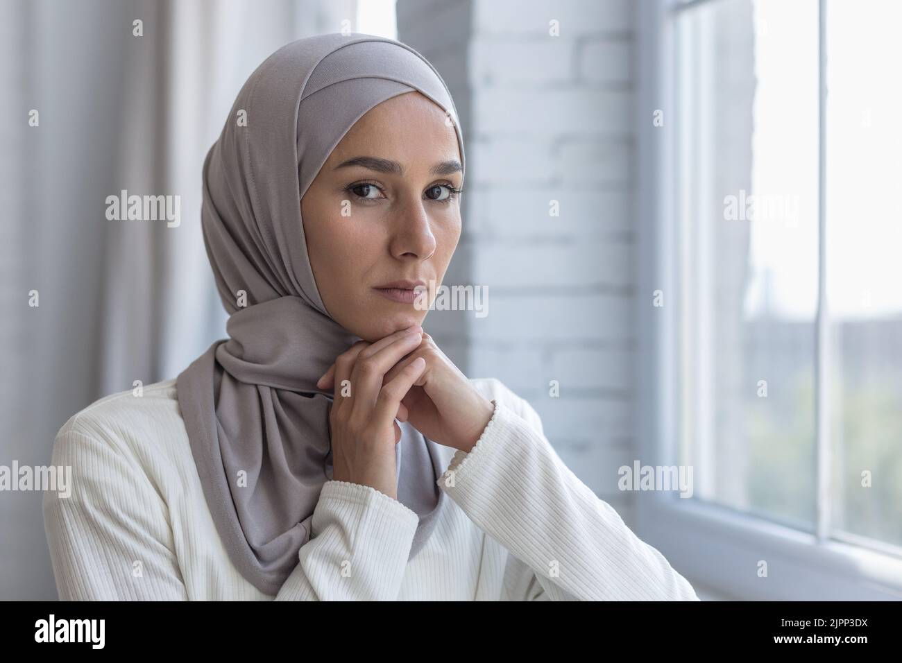 Ritratto di giovane bella donna musulmana vicino finestra a casa, donna araba in hijab guardando la macchina fotografica con concentrazione, primo piano foto Foto Stock