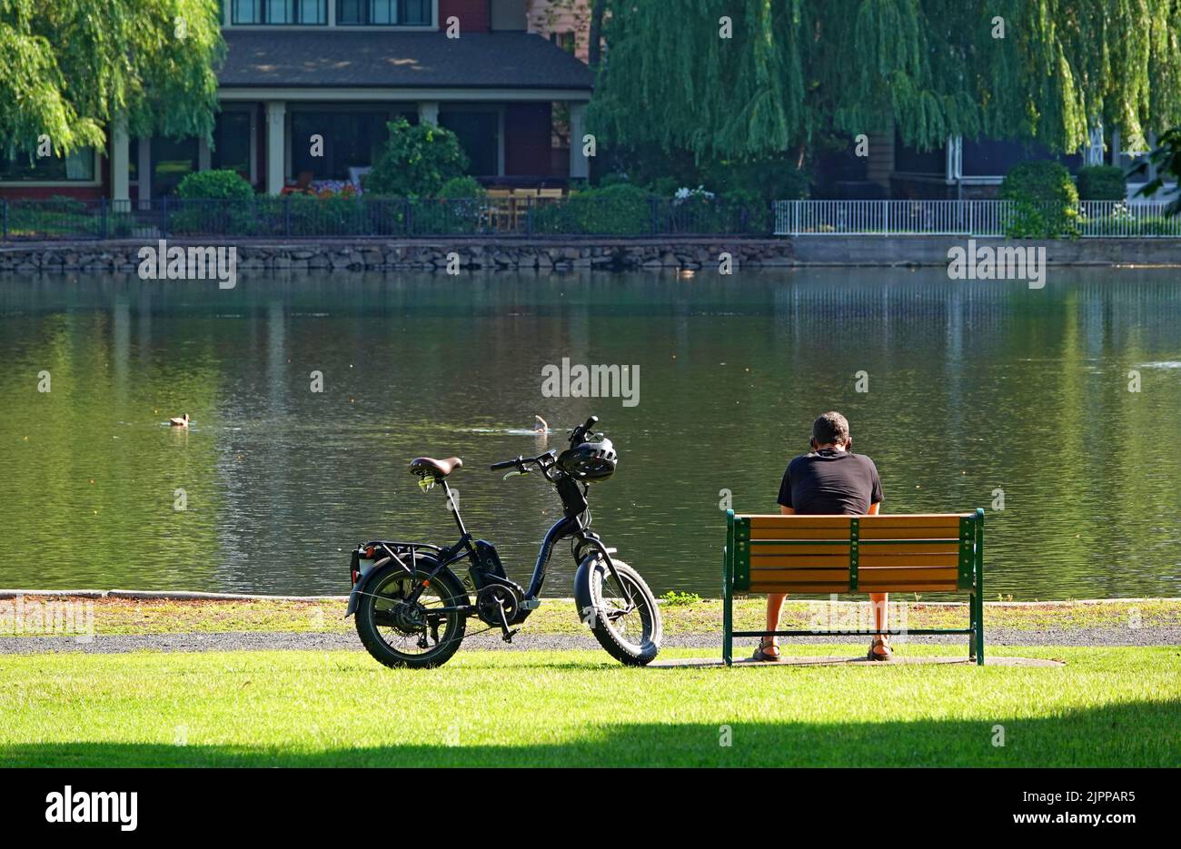Una bicicletta elettrica accanto a un ragazzo seduto su una panchina del parco lungo il fiume Deschutes a Bend, Oregon, durante l'estate. Drake Park Foto Stock