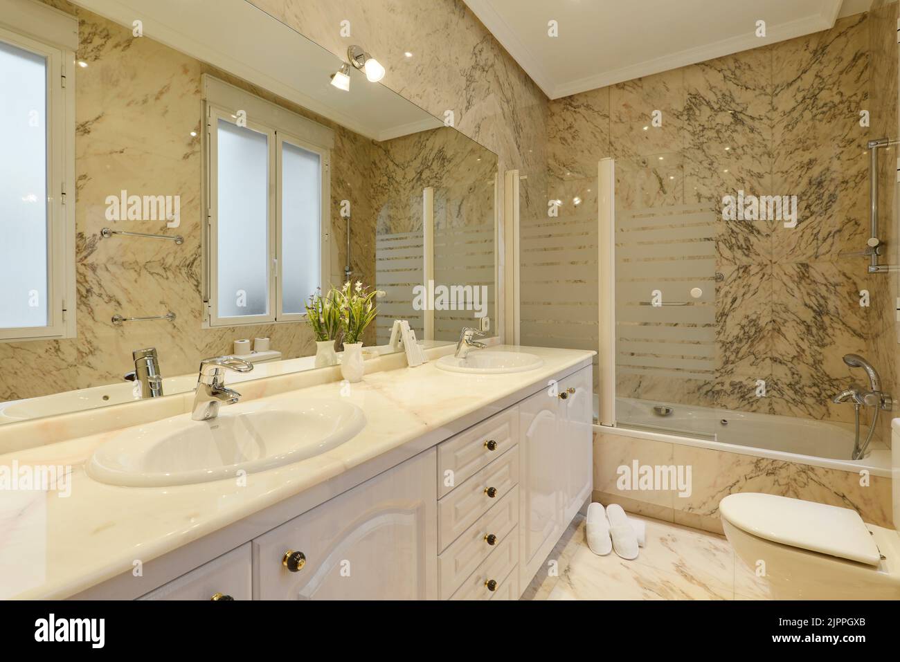 Bagno con lungo piano in marmo con due lavandini, grande specchio senza cornice alle pareti e piastrelle in marmo alle pareti e ai pavimenti Foto Stock