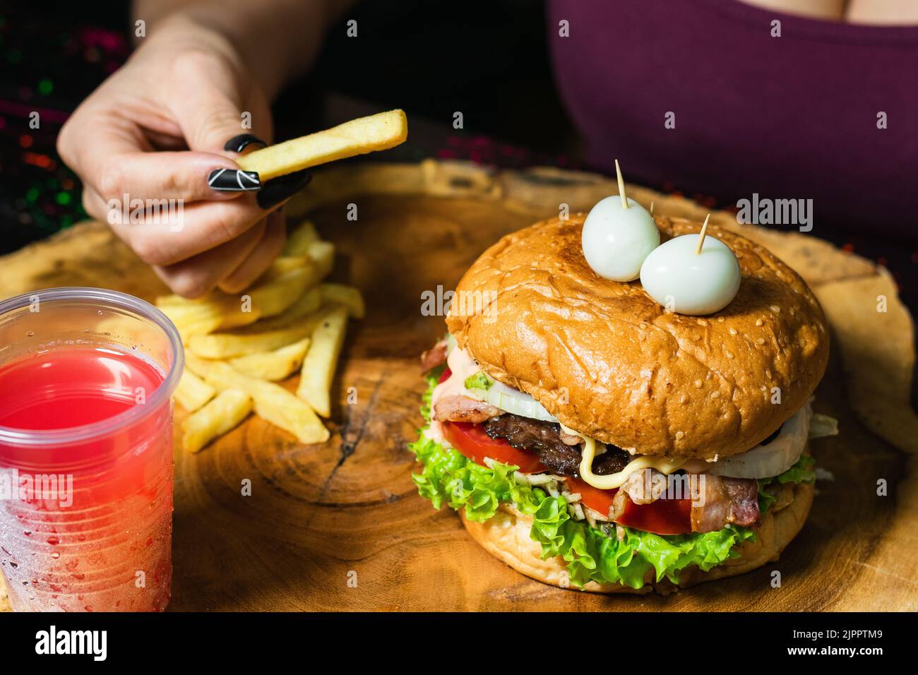 primo piano di una ragazza latina con il suo hamburger con patatine fritte e succo di frutta rossa, servito sul tavolo di legno, pronto per iniziare a mangiare. donna con una patata in Foto Stock