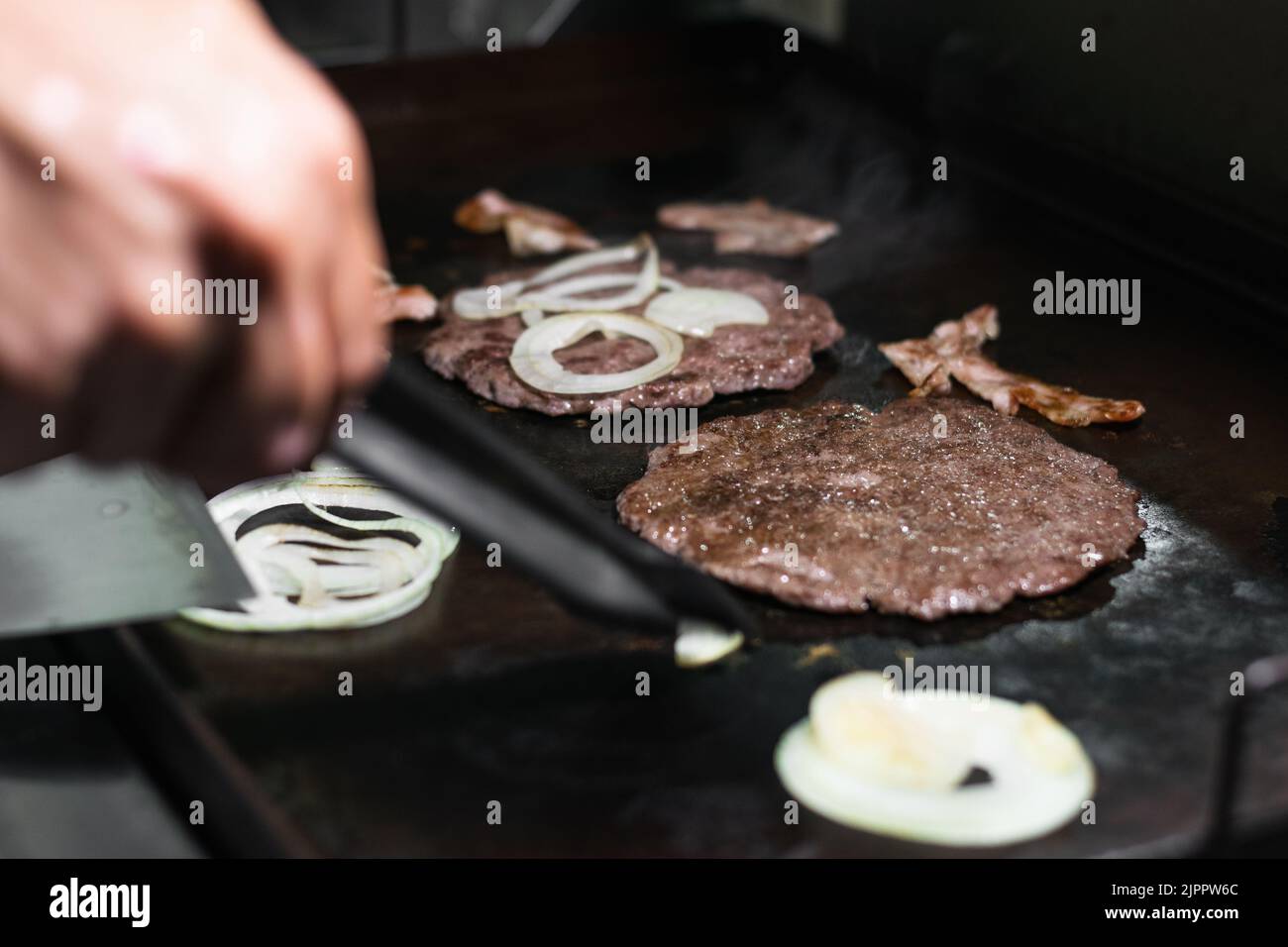 Dettaglio primo piano di un uomo che griglia due hamburger di carne macinata con cipolle e pancetta intorno a loro, cucinando in una griglia colombiana Street food o fast food Foto Stock