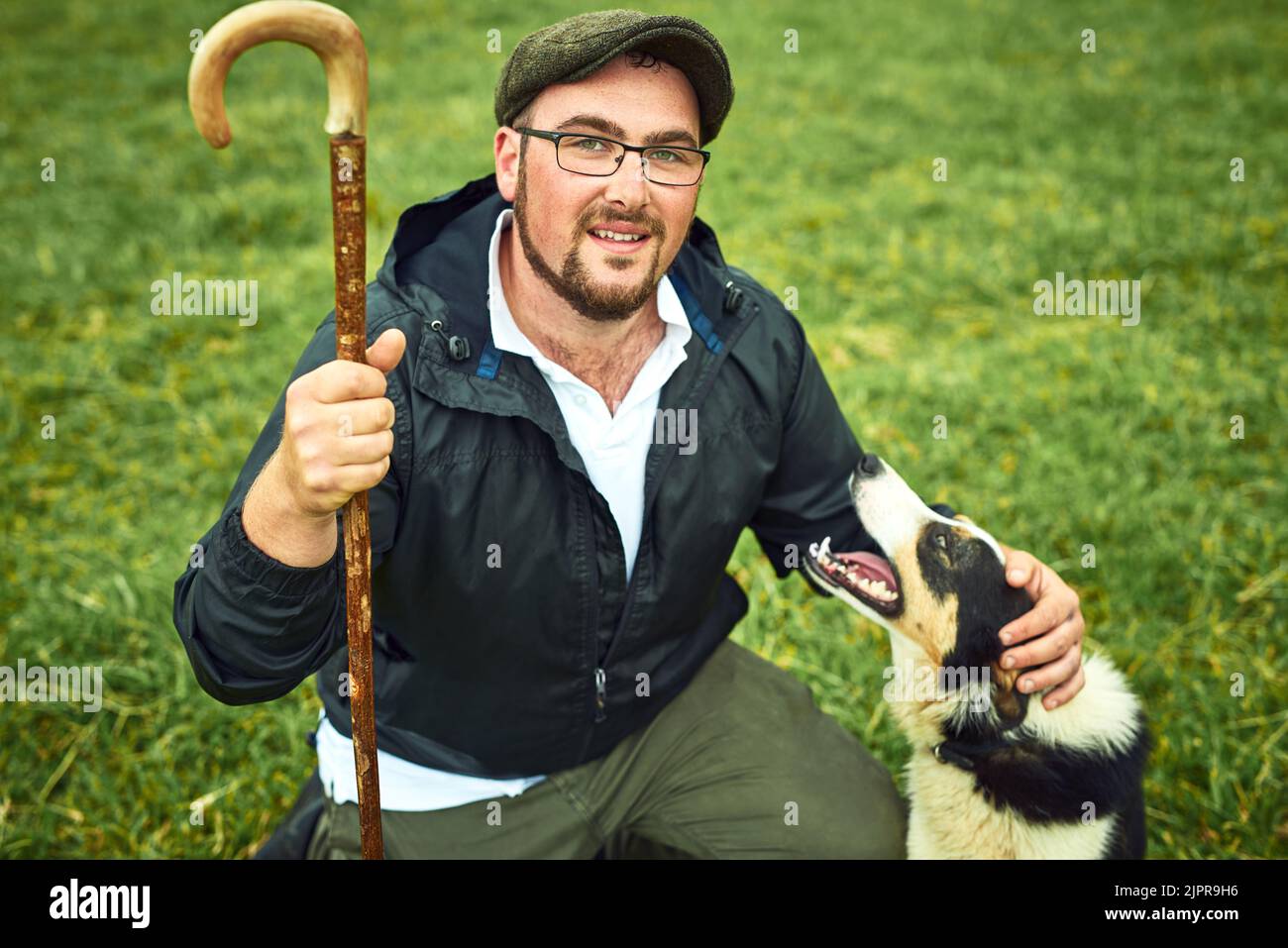 Oggi ci accingiamo ad allevare qualche pecora. Ritratto di un giovane contadino allegro e il suo cane che si rilassa su un campo verde in una fattoria durante il giorno. Foto Stock