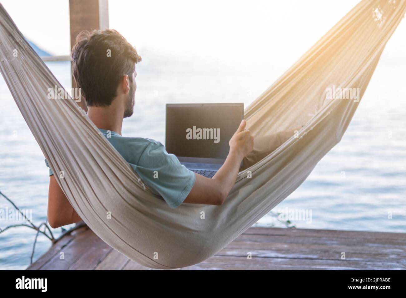 Giovane nomade digitale sdraiato su amaca con vista mare lavorare a distanza su internet al tramonto - viaggiare con un computer - sogno online di concetto di lavoro - se Foto Stock