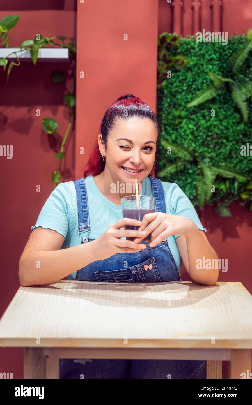 Donna che sorride e si strinca alla macchina fotografica mentre beve un frullato in una caffetteria. Concetto di stile di vita sano e bevande. Foto Stock