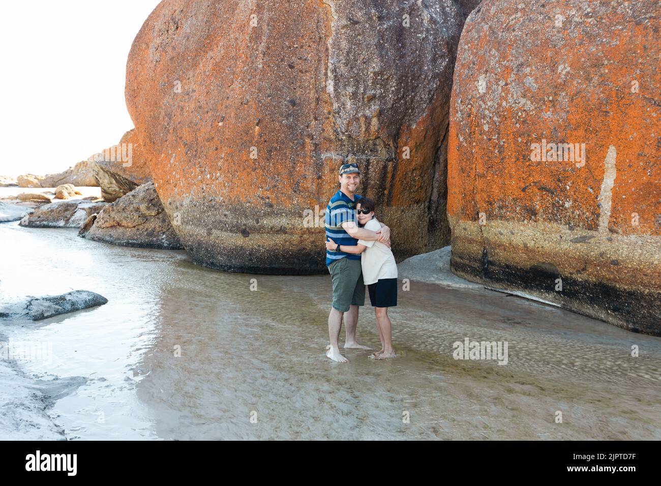 Un padre e un figlio che indossano occhiali da sole e un cappello accanto a rocce di masso sulla spiaggia Squeaky nel promontorio South Gippsland Australia di Wilson Foto Stock