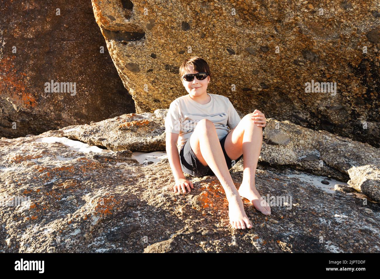 Un bambino che indossa occhiali da sole seduto su rocce di masso a Squeaky Beach nel promontorio di Wilson, South Gippsland Australia Foto Stock