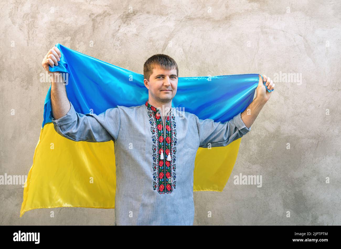 Il giovane tiene sopra la testa la bandiera blu-gialla dell'Ucraina. Uomo in camicia tradizionale ricamata sullo sfondo di pareti in cemento testurizzato. Vista frontale, spazio di copia. Giornata dell'indipendenza dell'Ucraina Foto Stock
