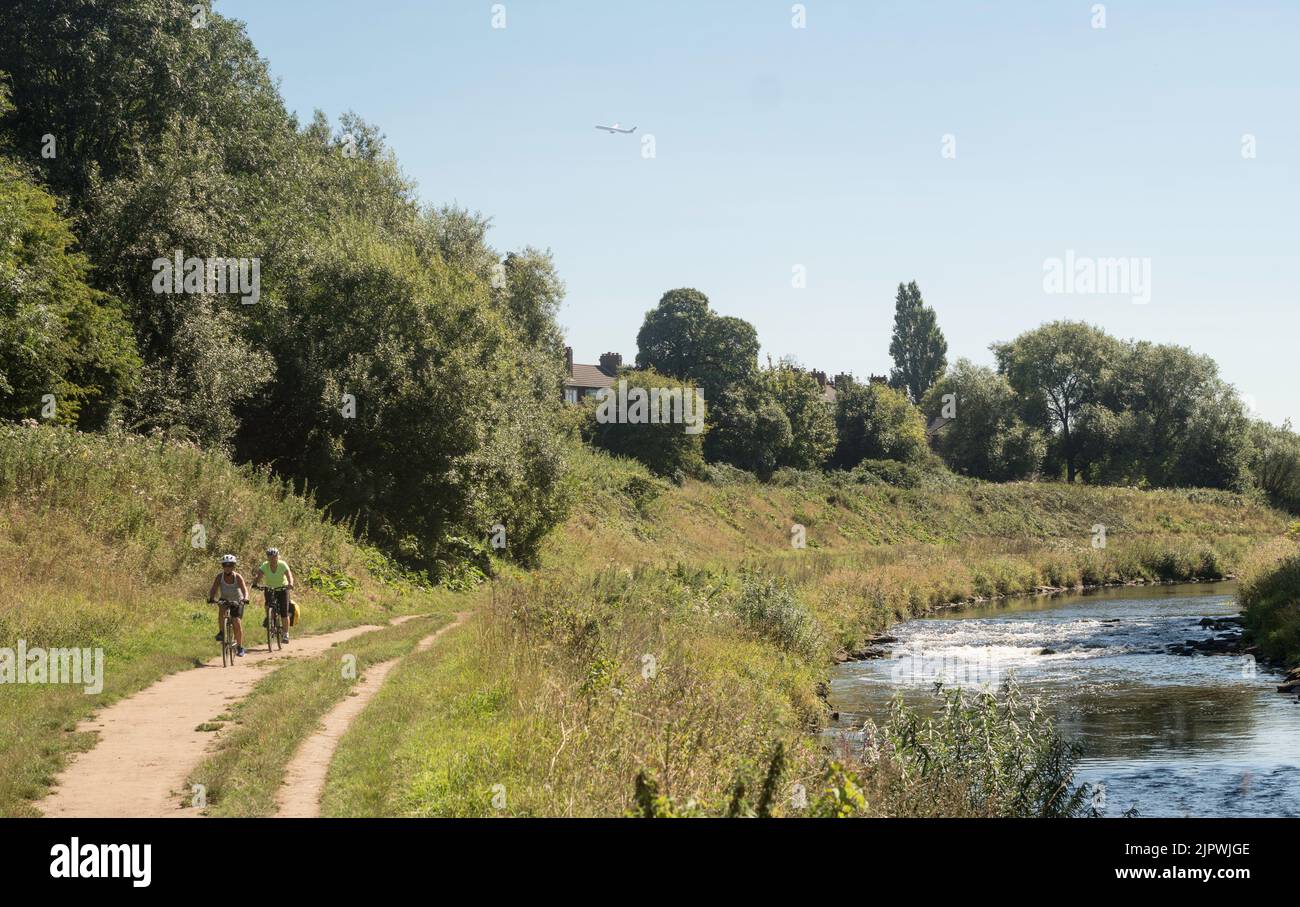 Due ciclisti femminili che cavalcano lungo un sentiero adiacente al fiume Mersey a Chorlton, Manchester come un aereo vola sopra, Inghilterra, Regno Unito Foto Stock