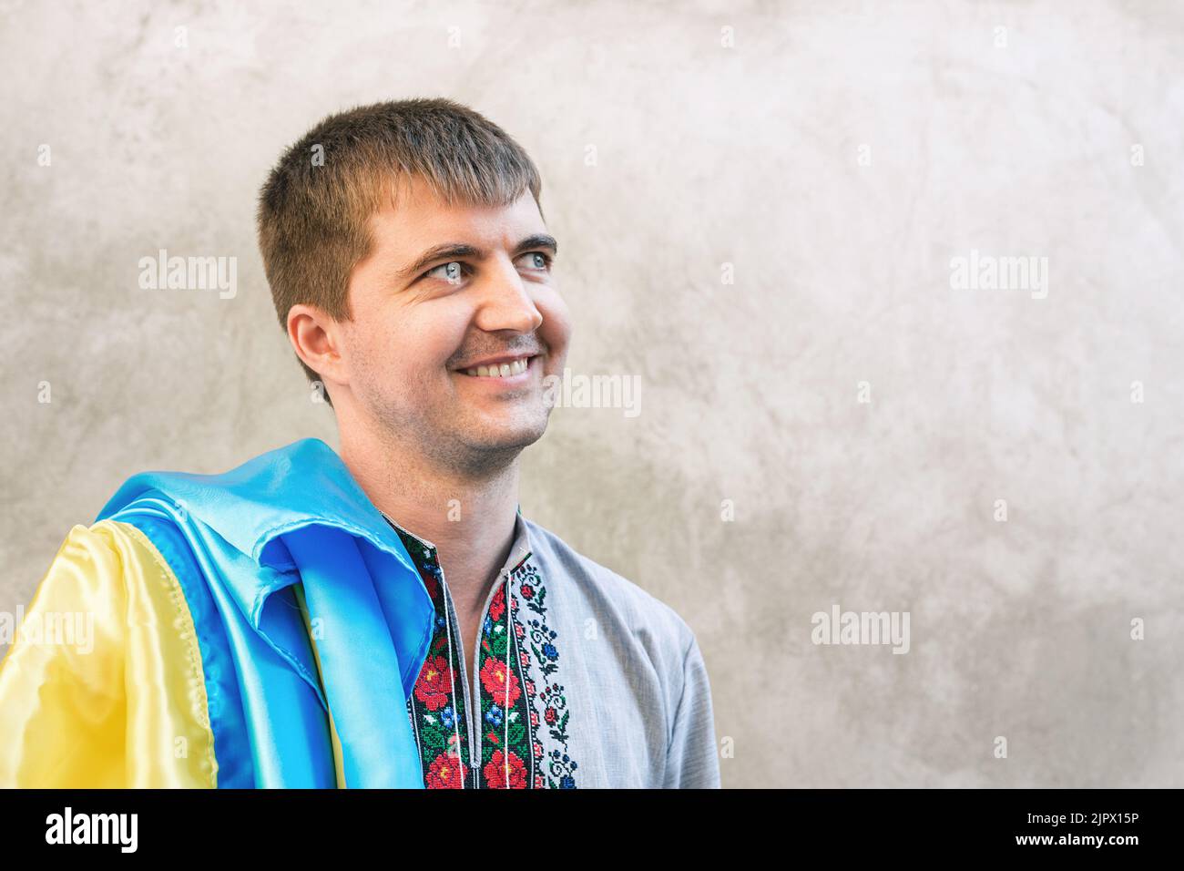 Primo piano ritratto di un giovane con bandiera blu e gialla dell'Ucraina. Uomo in camicia tradizionale ricamata sullo sfondo di parete grigia in città. Vista frontale, Copia spazio. Giornata dell'indipendenza dell'Ucraina Foto Stock