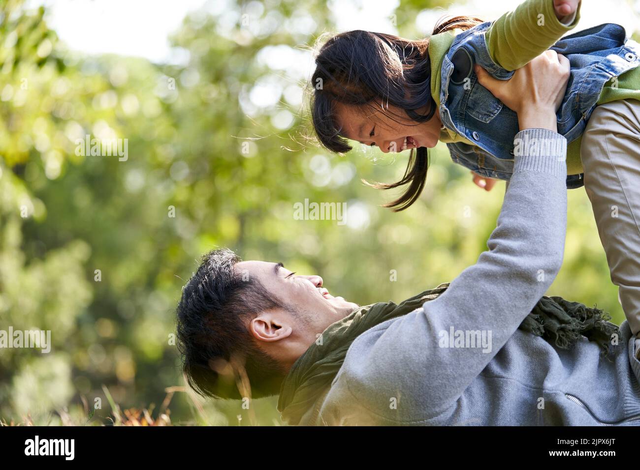 padre asiatico che si trova sulla schiena su erba avendo una buona volta sollevare figlia in aria all'aperto nel parco della città Foto Stock