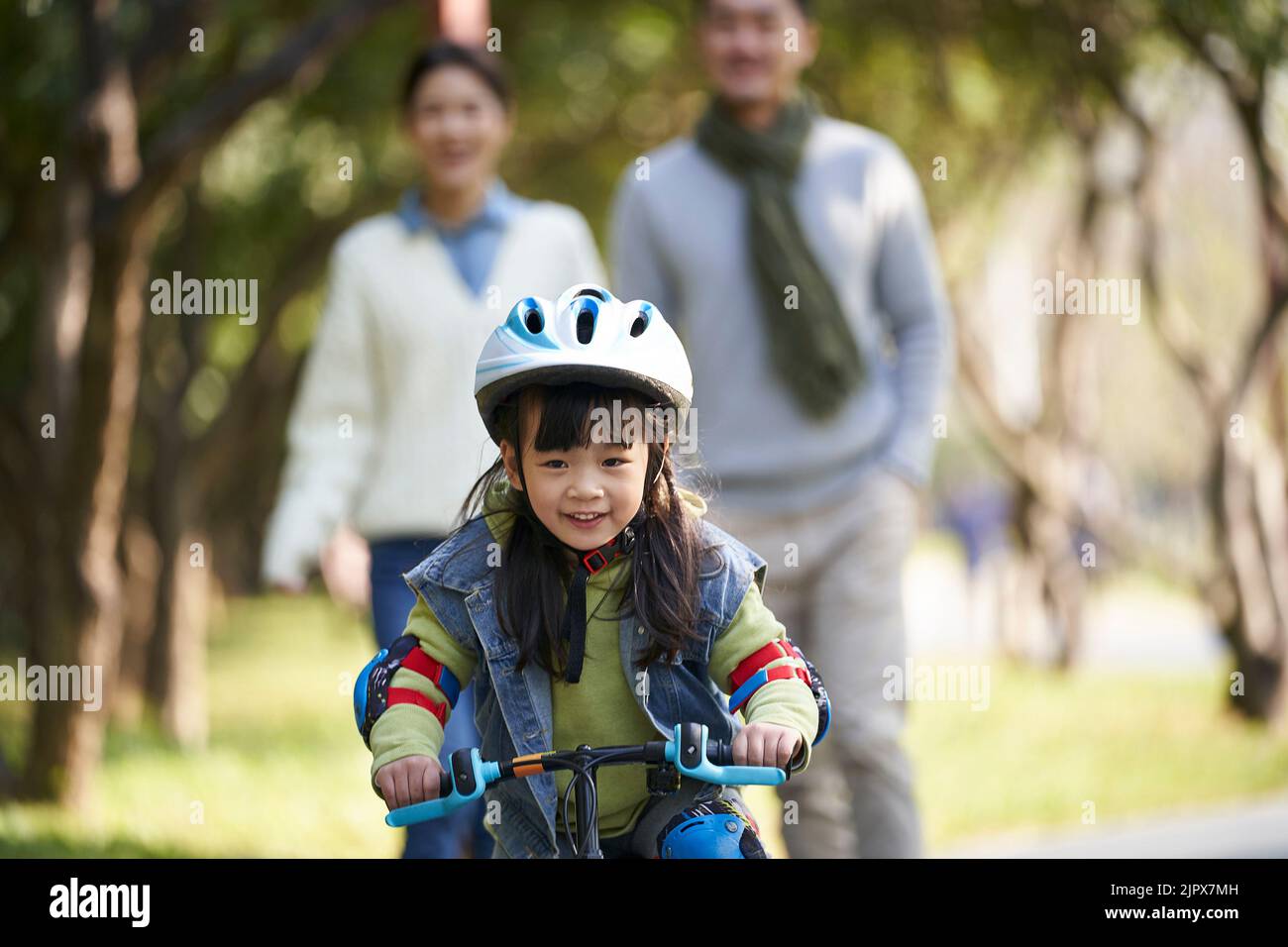 bambina asiatica con casco e completo di protezione in bicicletta nel parco della città con i genitori che guardano da dietro Foto Stock