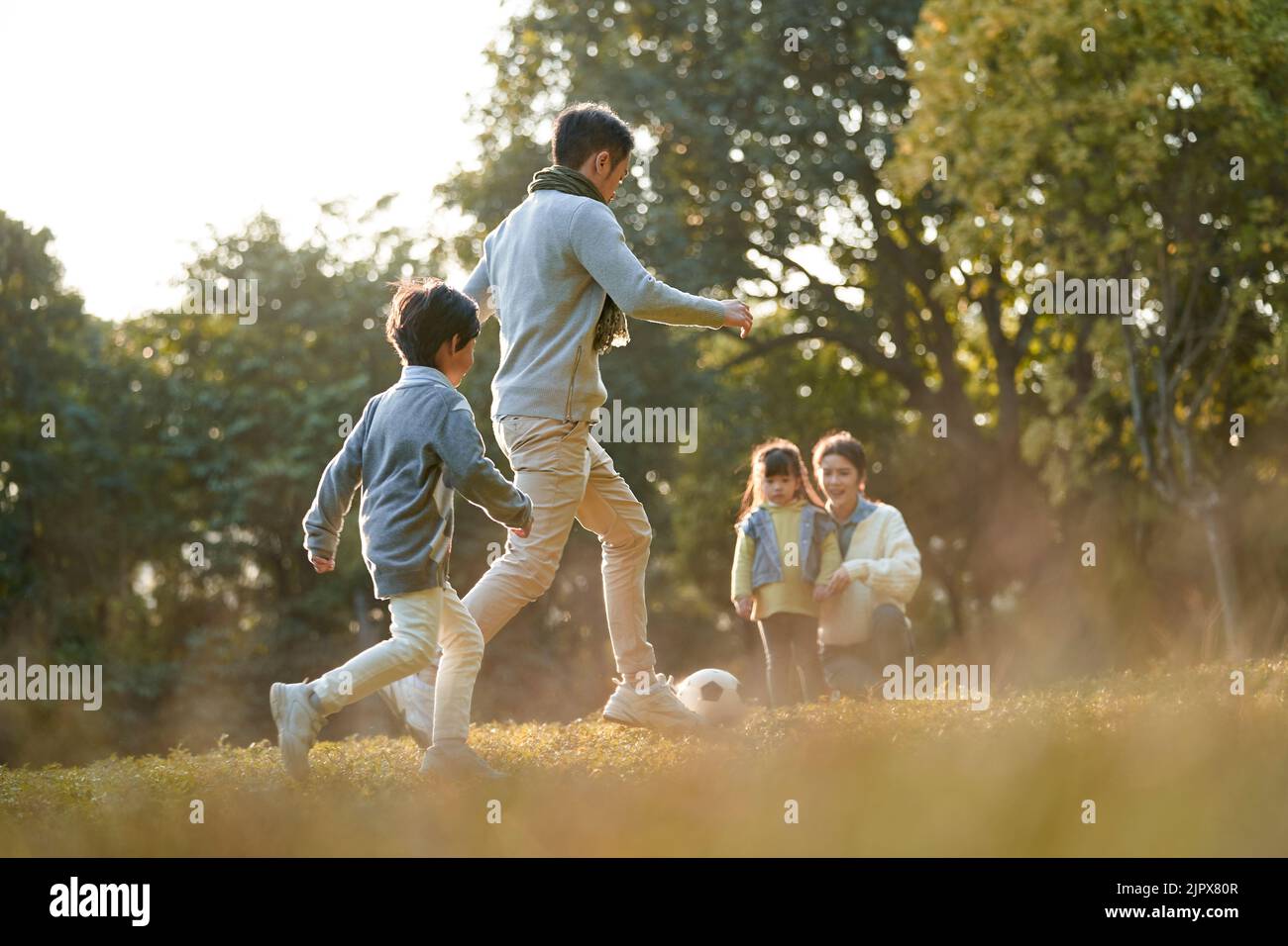 il padre e il figlio asiatico giocano a calcio all'aperto nel parco mentre la madre e la figlia guardano al fianco Foto Stock